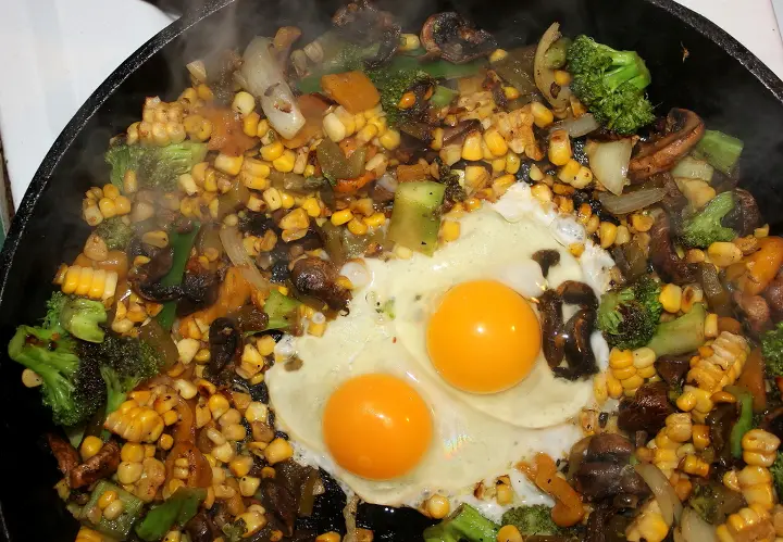 Egg skillet with two sunny side up eggs cooking atop a steaming pan of crisp sauteed veggies
